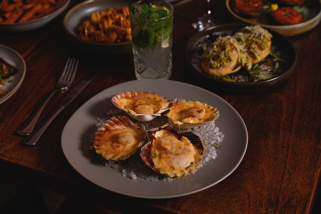 Cheesy scallops in their shells, browned on top and resting on rock salt, with a fresh mojito, sweet potato fries and herb-topped empanadas just visible in the background.