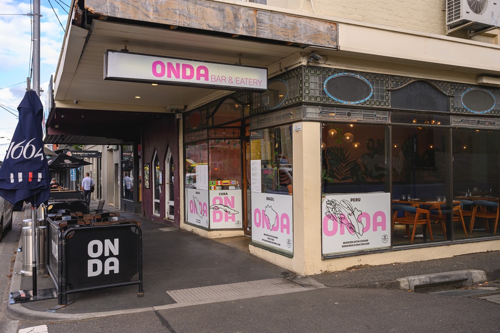 ONDA’s corner storefront at 312 Brunswick Street, Fitzroy — pink ONDA Bar & Eatery signage, country posters for Mexico, Cuba, Brazil and Peru, curved heritage windows and warm interior glow.