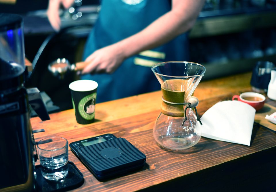 A barista at a timber cafe counter with a takeaway cup, scales and a pour-over