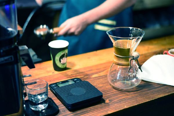 A barista at a timber cafe counter with a takeaway cup, scales and a pour-over